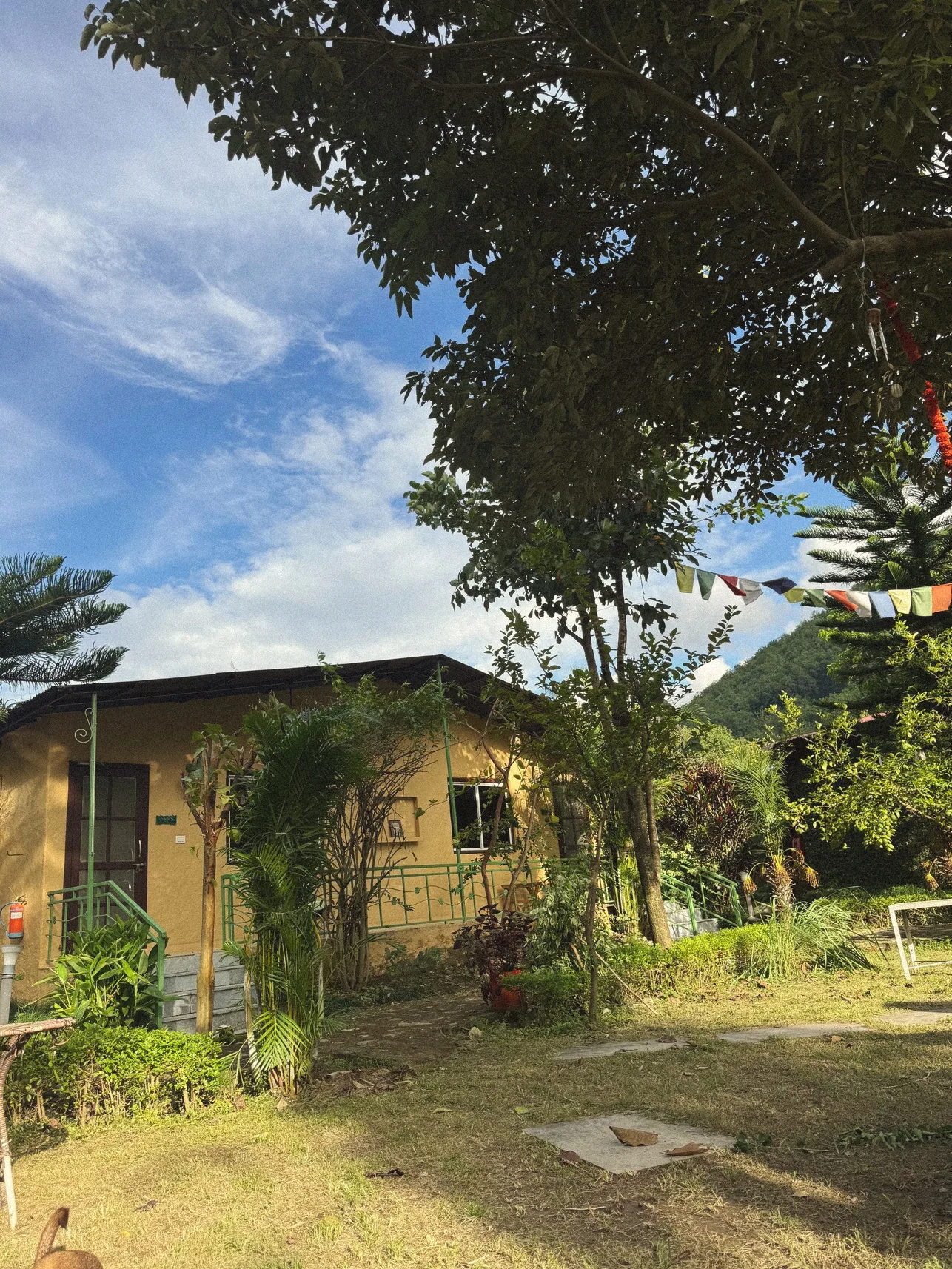 Ochre cottage in a garden with prayer flags against a blue sky and forested hill